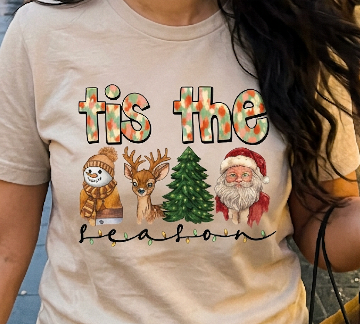 Close-up of sand Tis the Season Christmas shirt showing colorful watercolor Christmas characters, holiday lights, and decorative seasonal typography.