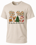 Sand-colored unisex Tis the Season Christmas t-shirt featuring watercolor Santa Claus, reindeer, snowman, Christmas tree, and festive holiday lettering.