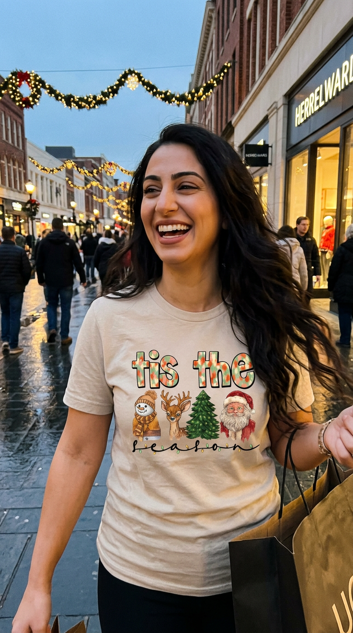 Woman wearing sand-colored Tis the Season Christmas graphic t-shirt while walking through a festive downtown shopping street decorated with holiday lights.