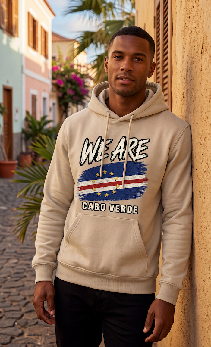 A man wearing a beige hoodie with a graphic print of the Cabo Verde flag and the text 'WE ARE CABO VERDE' stands against a warm-toned wall in an outdoor street setting.