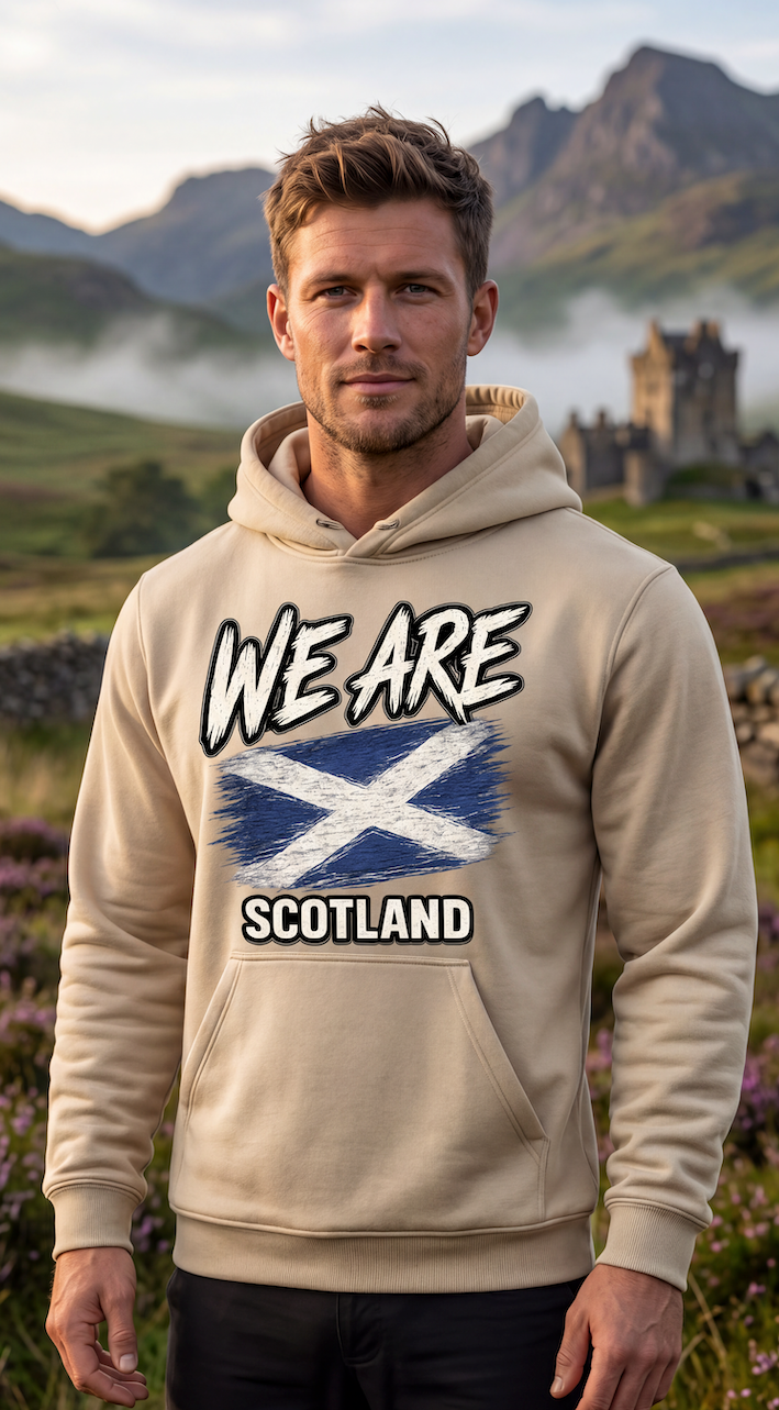 A man wearing a beige hoodie with a 'WE ARE SCOTLAND' graphic featuring the Scottish flag, standing in a scenic outdoor landscape with mountains and a castle in the background.