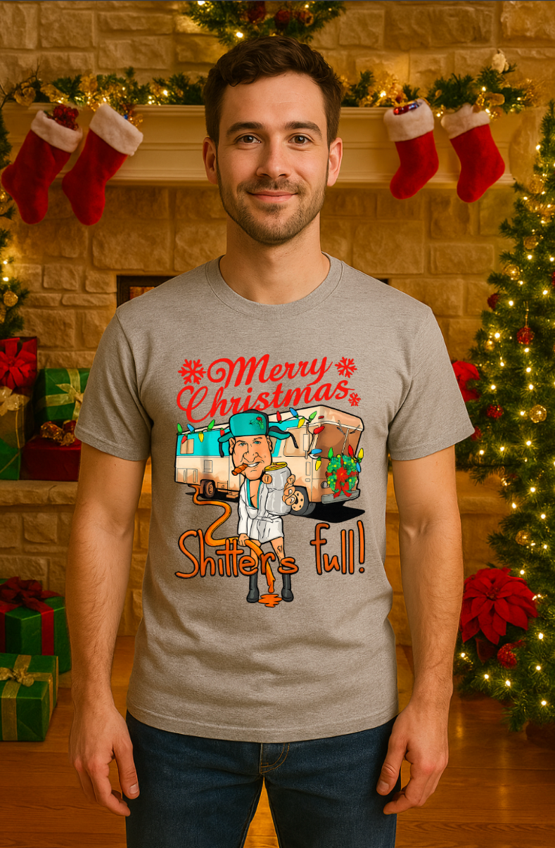 Smiling man wearing a gray “Merry Christmas, Shitter’s Full” graphic t-shirt, standing in a festive holiday room with a decorated fireplace, stockings, Christmas lights, and wrapped gifts.