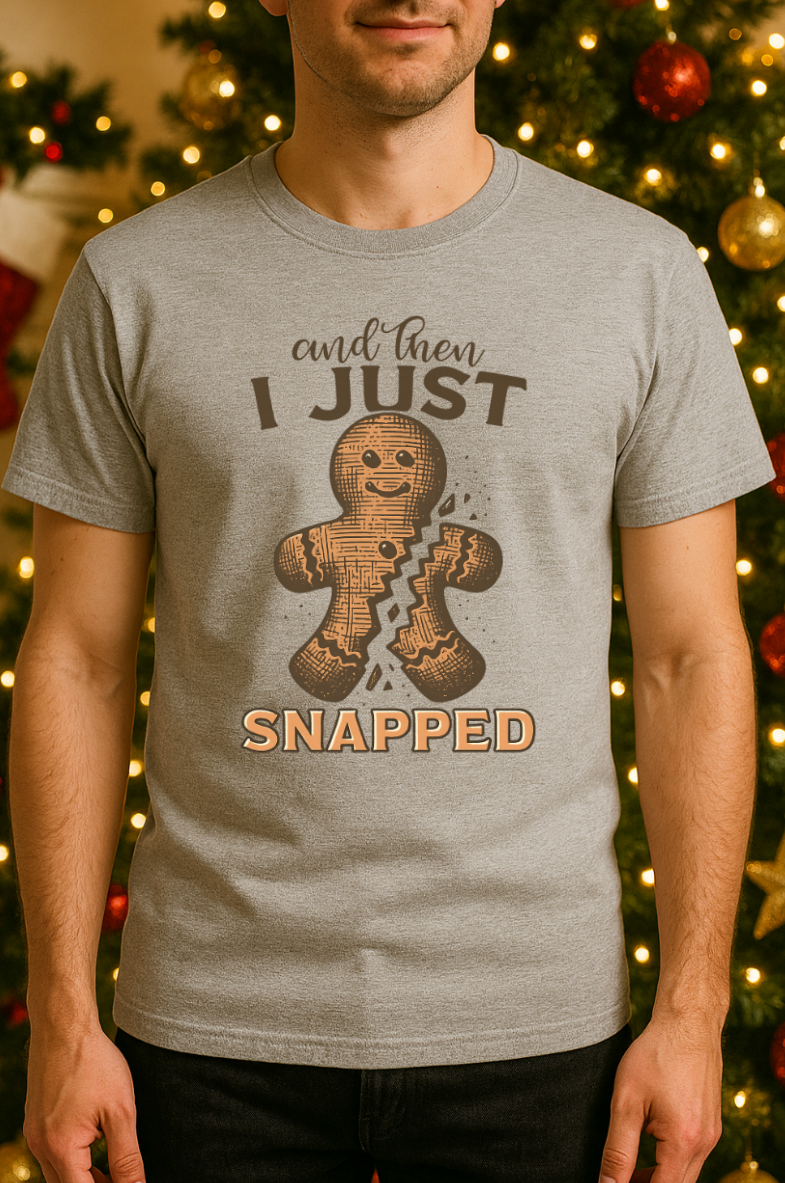 Man wearing a gray “And Then I Just Snapped” gingerbread Christmas shirt, standing in front of a decorated Christmas tree with holiday lights.
