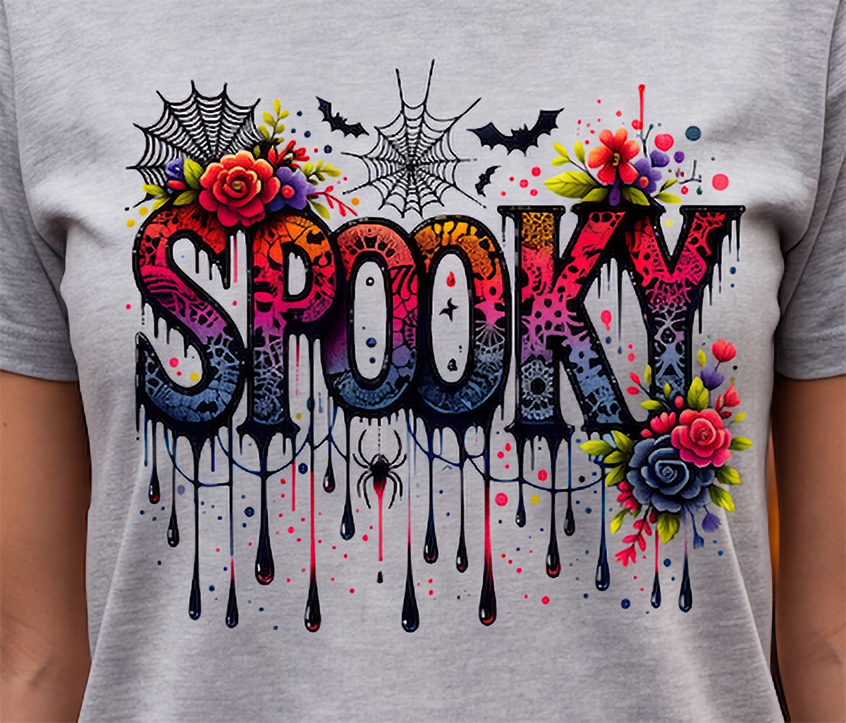 A close-up of a person wearing a light gray heather t-shirt with a vibrant, multicolor 'Spooky' graphic print featuring dripping letters, roses, bats, and spiderwebs.