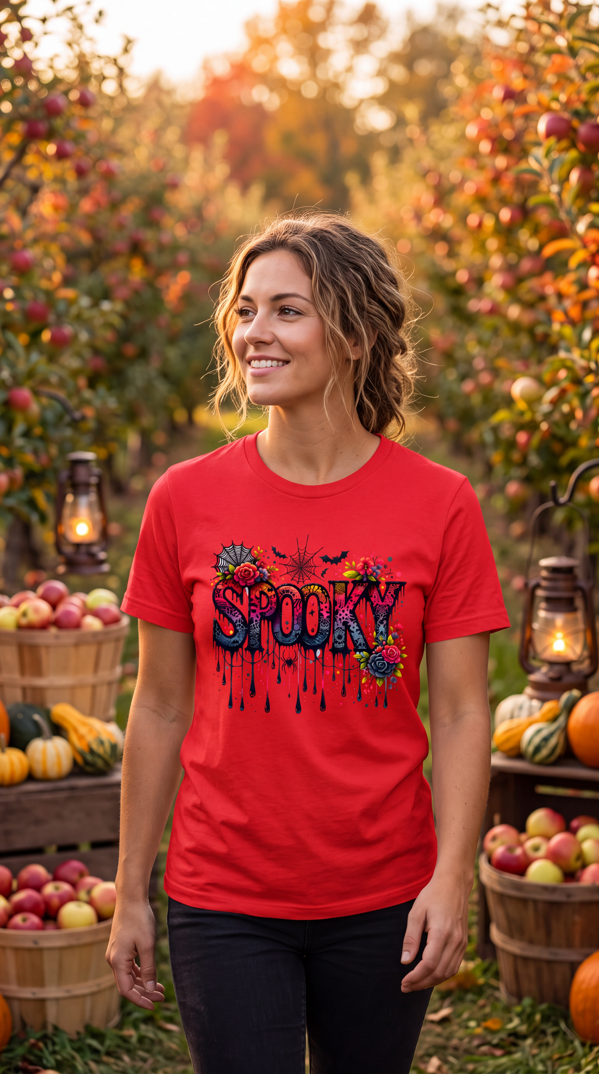 A woman stands in an autumn orchard wearing a bright red graphic T-shirt with the word 'SPOOKY' in colorful dripping letters. Baskets of apples and pumpkins, lanterns, and warm sunlight fill the background.