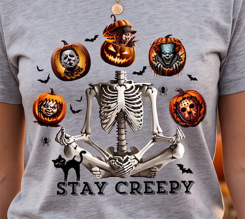 A close-up of a person wearing a heather gray t-shirt with a detailed Halloween graphic. The design features a central white skeleton meditating, surrounded by five carved pumpkins with horror character faces (Michael Myers, a zombie, a skull-faced figure, a clown pumpkin, and a pumpkin skull), small black bats, a black cat, and the text 'STAY CREEPY' printed at the bottom.