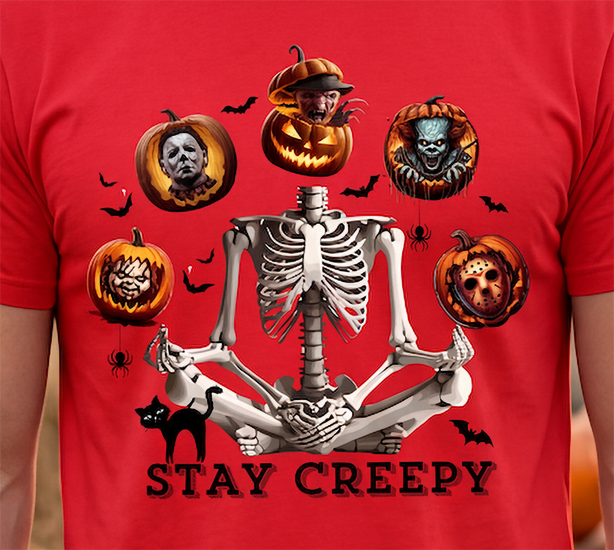 A red short-sleeve t-shirt featuring a graphic of a skeleton meditating, surrounded by various horror-themed jack-o'-lanterns, a black cat, bats, and the text 'STAY CREEPY'.