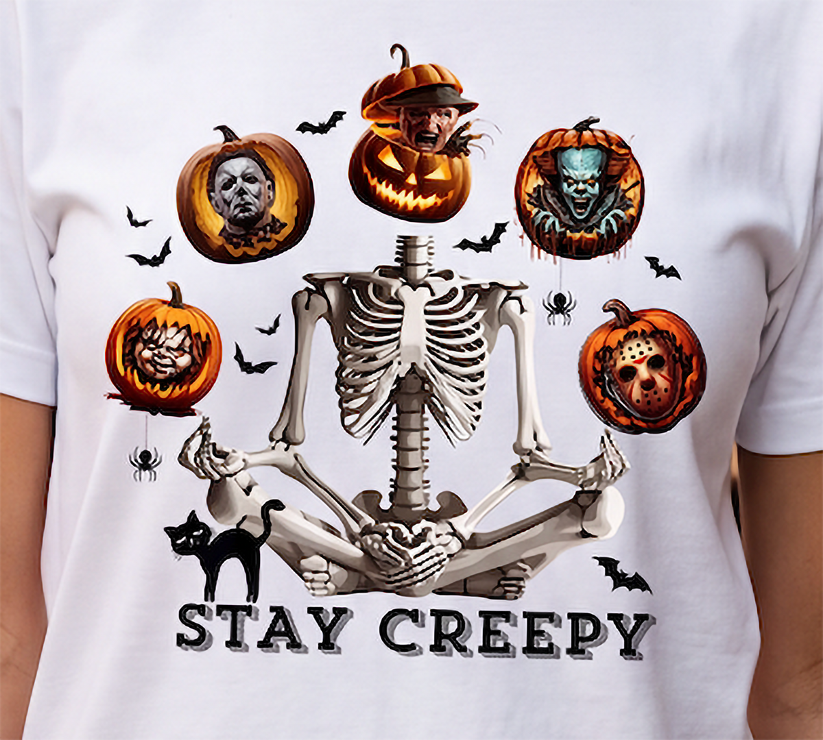A white short-sleeve T-shirt featuring a large Halloween graphic of a meditating skeleton surrounded by spooky jack-o'-lanterns, bats, spiders, a black cat, and the text 'STAY CREEPY'.