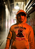 Man wearing orange “Stay Sharp” graphic hoodie in a graffiti alley streetwear setting