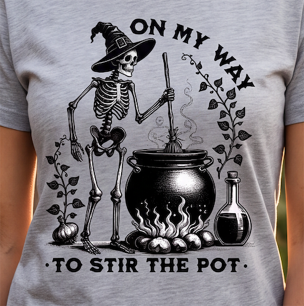 A close-up of a light heather gray t-shirt featuring a black graphic print of a witch skeleton stirring a cauldron with the text 'ON MY WAY TO STIR THE POT'.