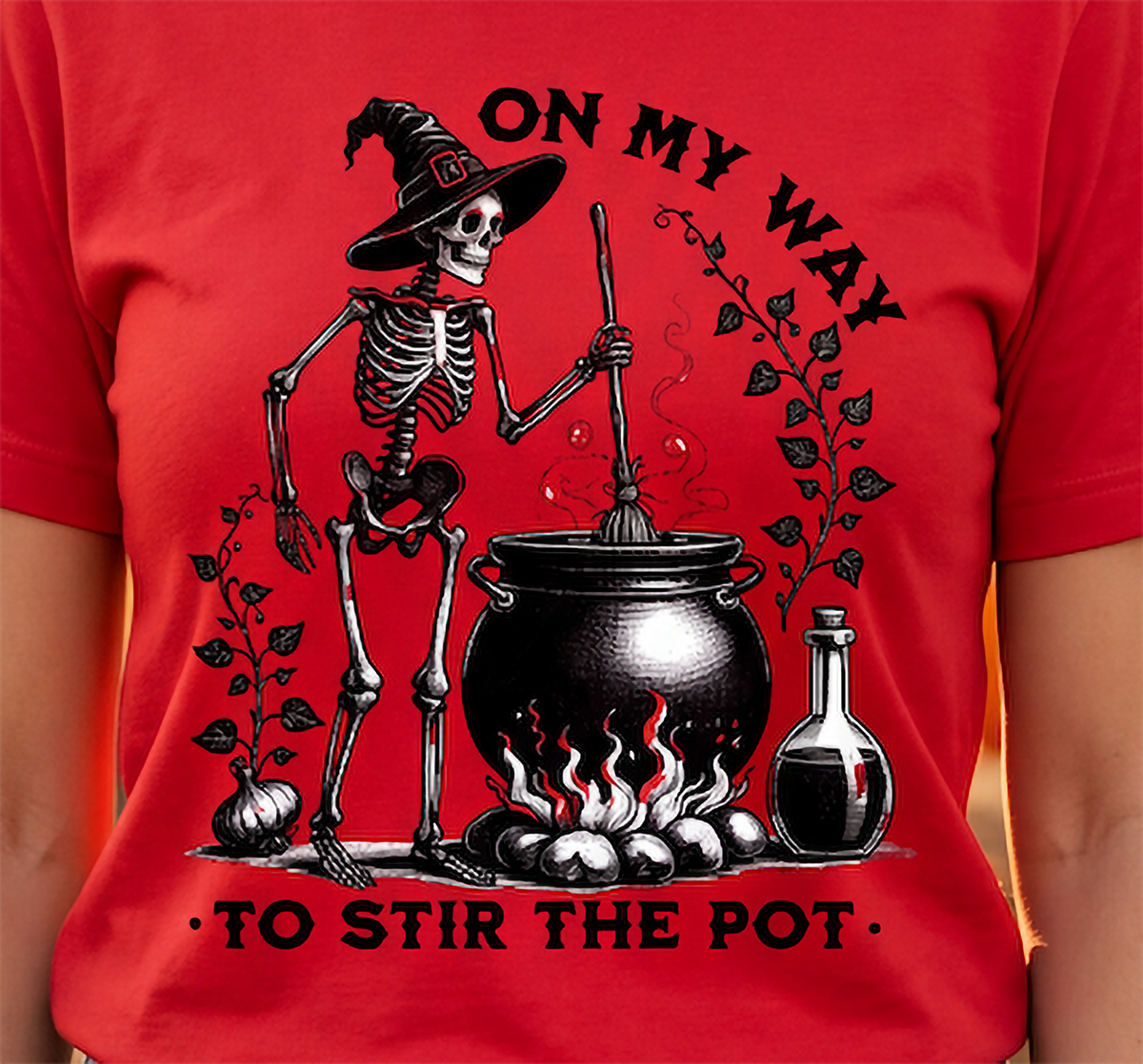 A red short-sleeve t-shirt featuring a black and white graphic of a skeleton wizard stirring a cauldron with the text 'ON MY WAY TO STIR THE POT'. The t-shirt is shown on a model, with the chest and arms visible.
