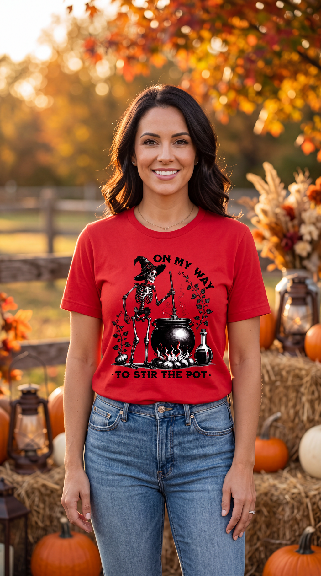 A smiling woman stands outdoors in an autumn yard wearing a bright red graphic T-shirt with a witch, cauldron, and the text 'ON MY WAY TO STIR THE POT.' She is also wearing blue jeans, with pumpkins, hay bales, lanterns, and fall foliage in the background.