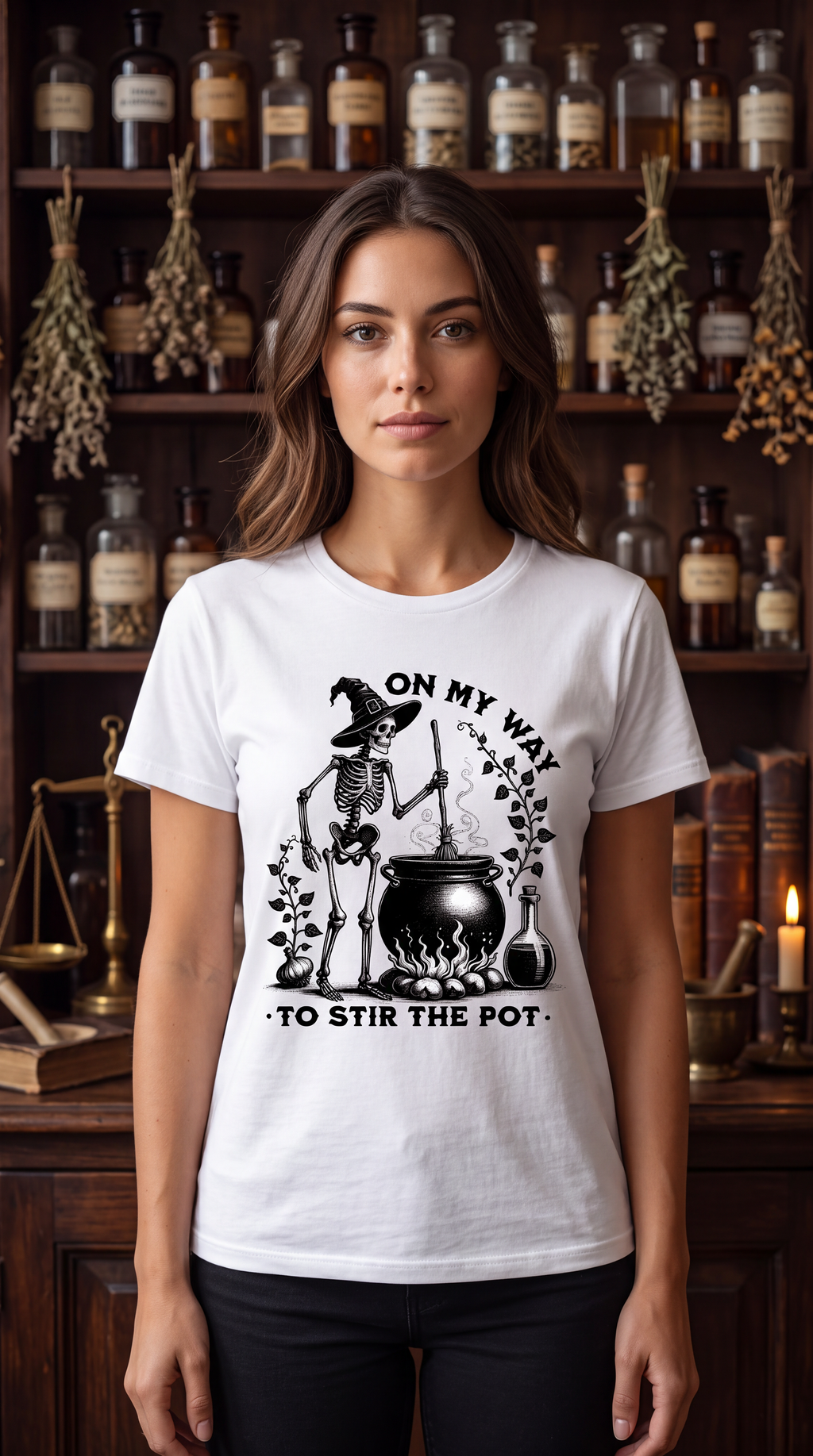 A woman stands in front of a dark wooden shelf filled with glass apothecary bottles and dried herbs, wearing a white graphic T-shirt with a black skeleton cauldron design and the text 'ON MY WAY TO STIR THE POT'.