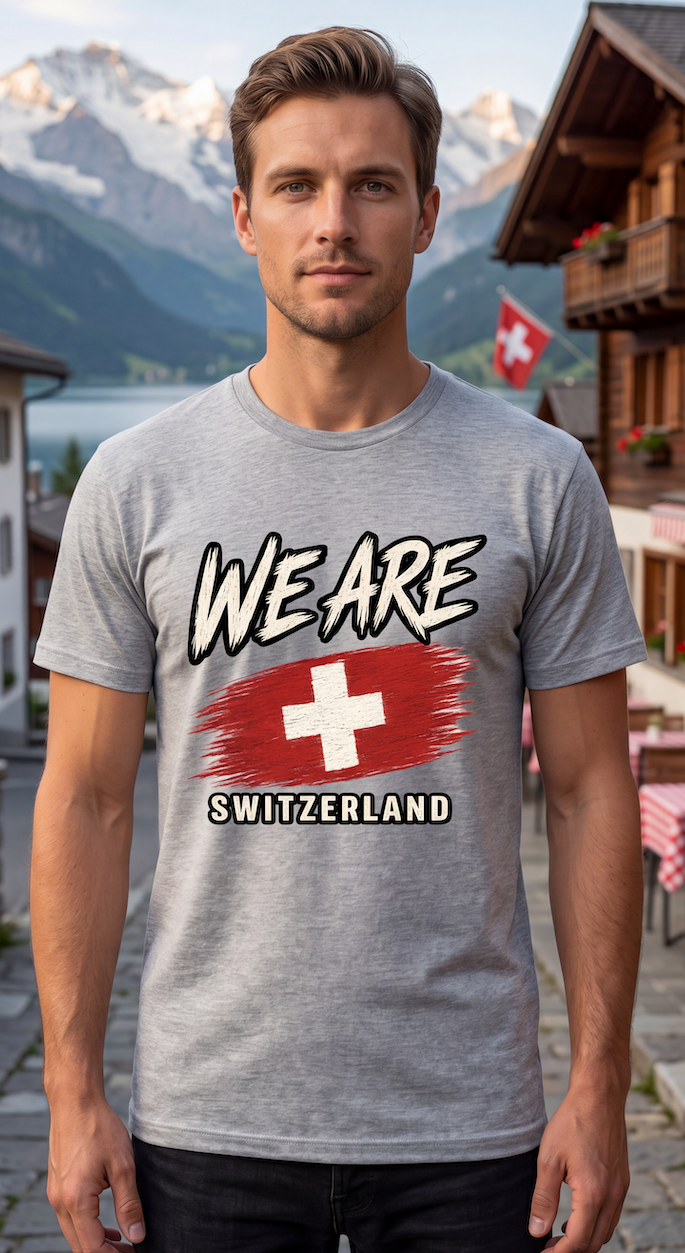 A man wearing a heather gray t-shirt with a graphic that reads 'WE ARE SWITZERLAND' above and below a red and white Swiss flag design. The background shows a scenic mountain village with Swiss flags.