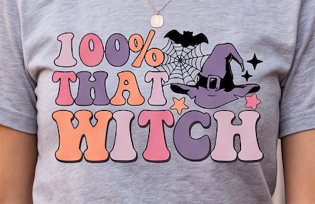 A close-up of a person wearing a heather gray t-shirt with a colorful graphic that reads '100% THAT WITCH' in bubble letters, featuring a purple witch hat, spiderweb, and stars.