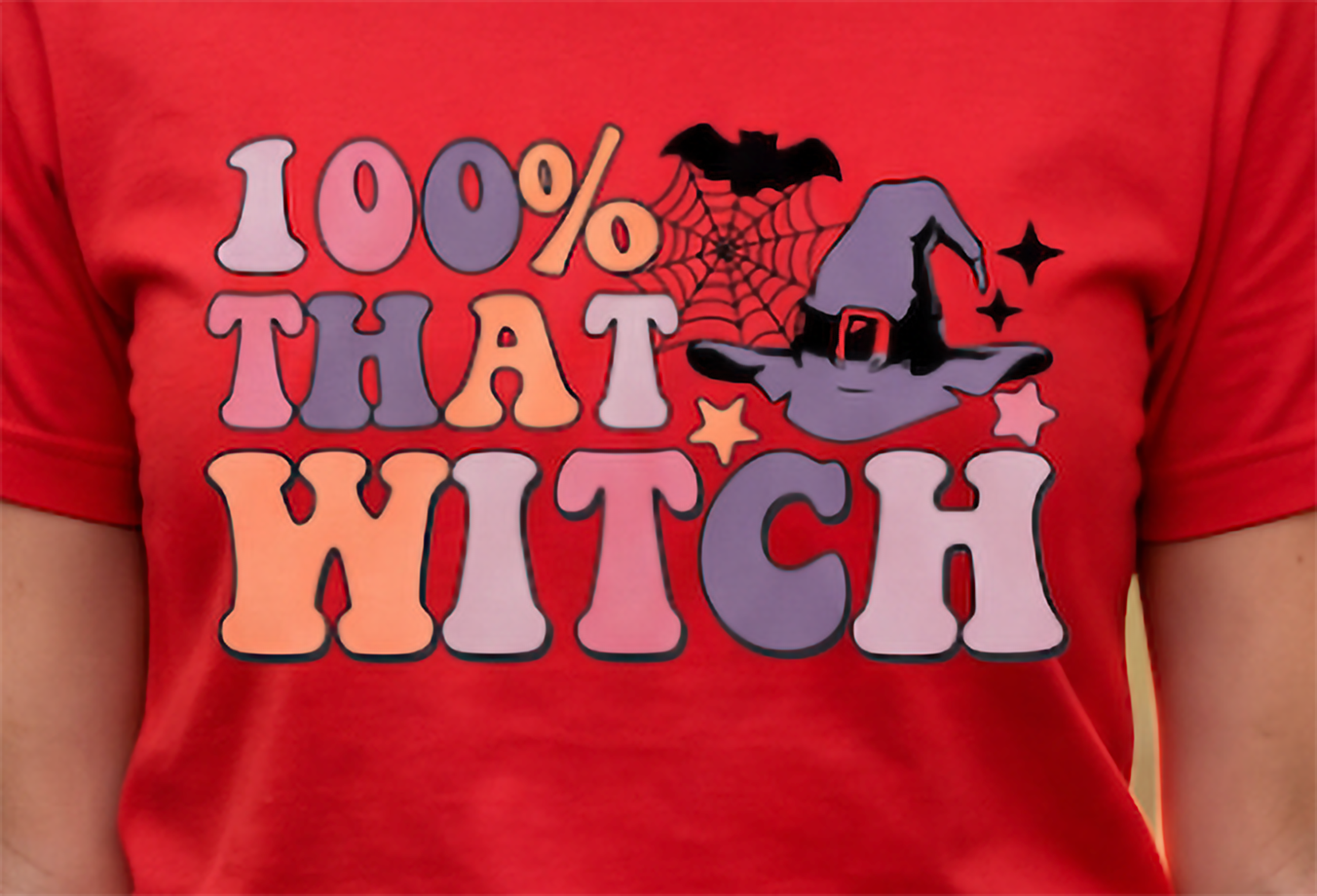 A close-up of a red short-sleeve T-shirt worn by a person, featuring a colorful retro-style graphic that reads '100% THAT WITCH' with a witch hat, spiderweb, bat, and stars.