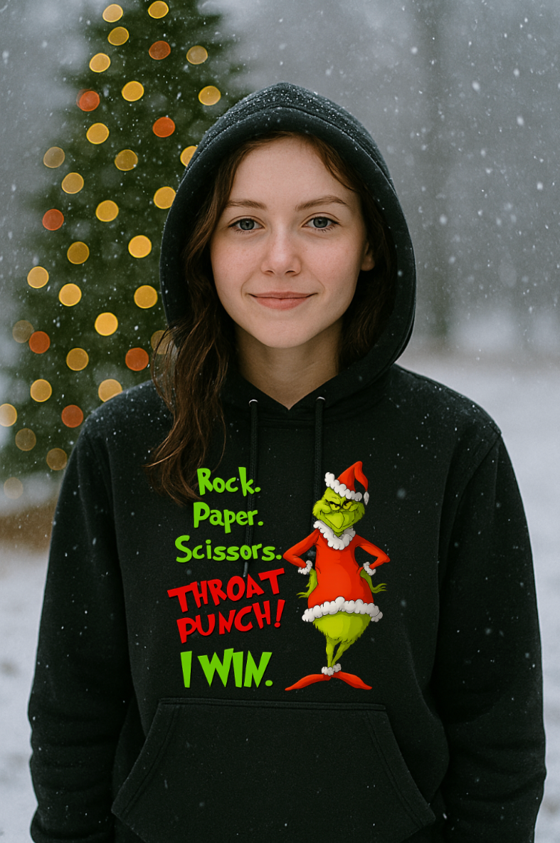 Woman wearing a black Christmas hoodie with a funny Rock Paper Scissors Throat Punch design, standing outside in falling snow with a decorated Christmas tree behind her.