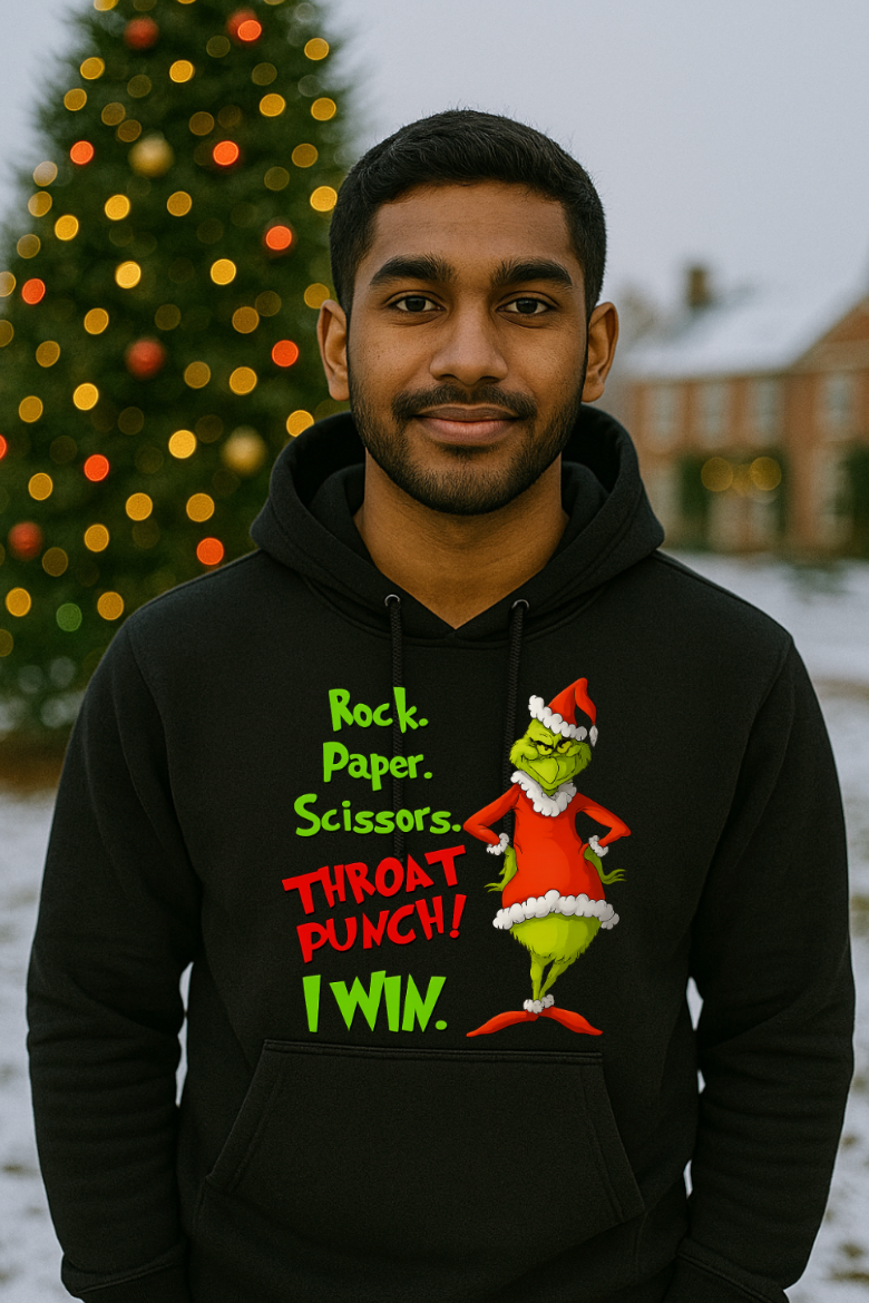 Man wearing a black Christmas hoodie with a funny Rock Paper Scissors Throat Punch design, standing outdoors in front of a decorated Christmas tree in a snowy setting.