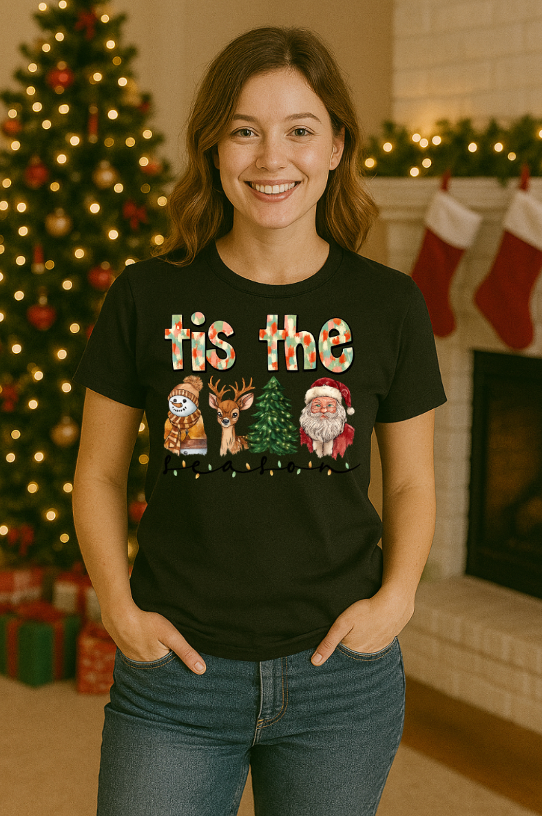 Woman wearing a black “’tis the season” Christmas graphic tee featuring a snowman, reindeer, Christmas tree, and Santa, standing in a festive living room with a decorated Christmas tree and stockings.