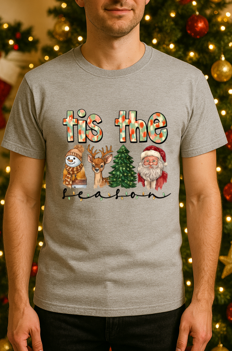 Man wearing a gray “’tis the season” Christmas graphic tee featuring a snowman, reindeer, Christmas tree, and Santa, standing in front of a decorated Christmas tree with lights and ornam