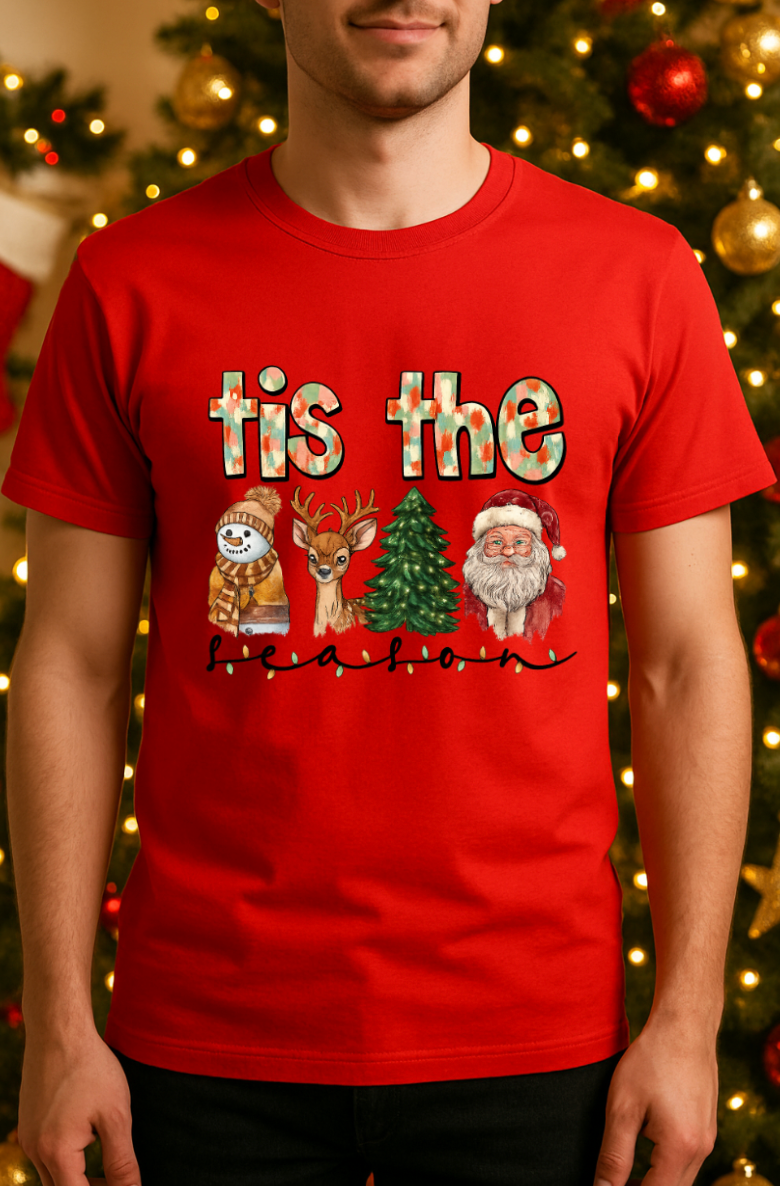 Man wearing a red “’tis the season” Christmas graphic tee featuring a snowman, reindeer, Christmas tree, and Santa, standing in front of a decorated Christmas tree with lights and ornaments.