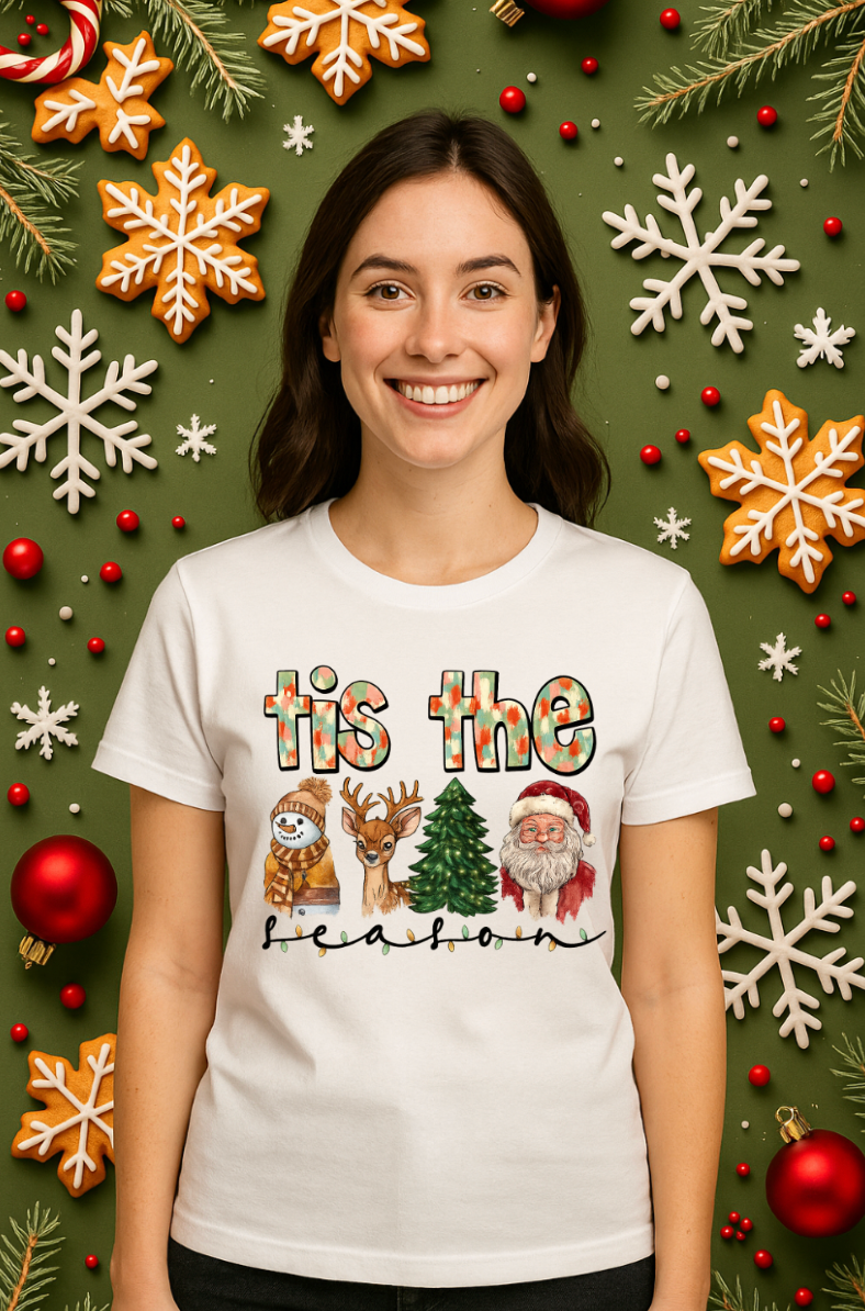 Smiling woman wearing a white “’tis the season” Christmas graphic tee with a snowman, reindeer, Christmas tree, and Santa, standing in front of a festive green background decorated with ornaments and snowflakes.