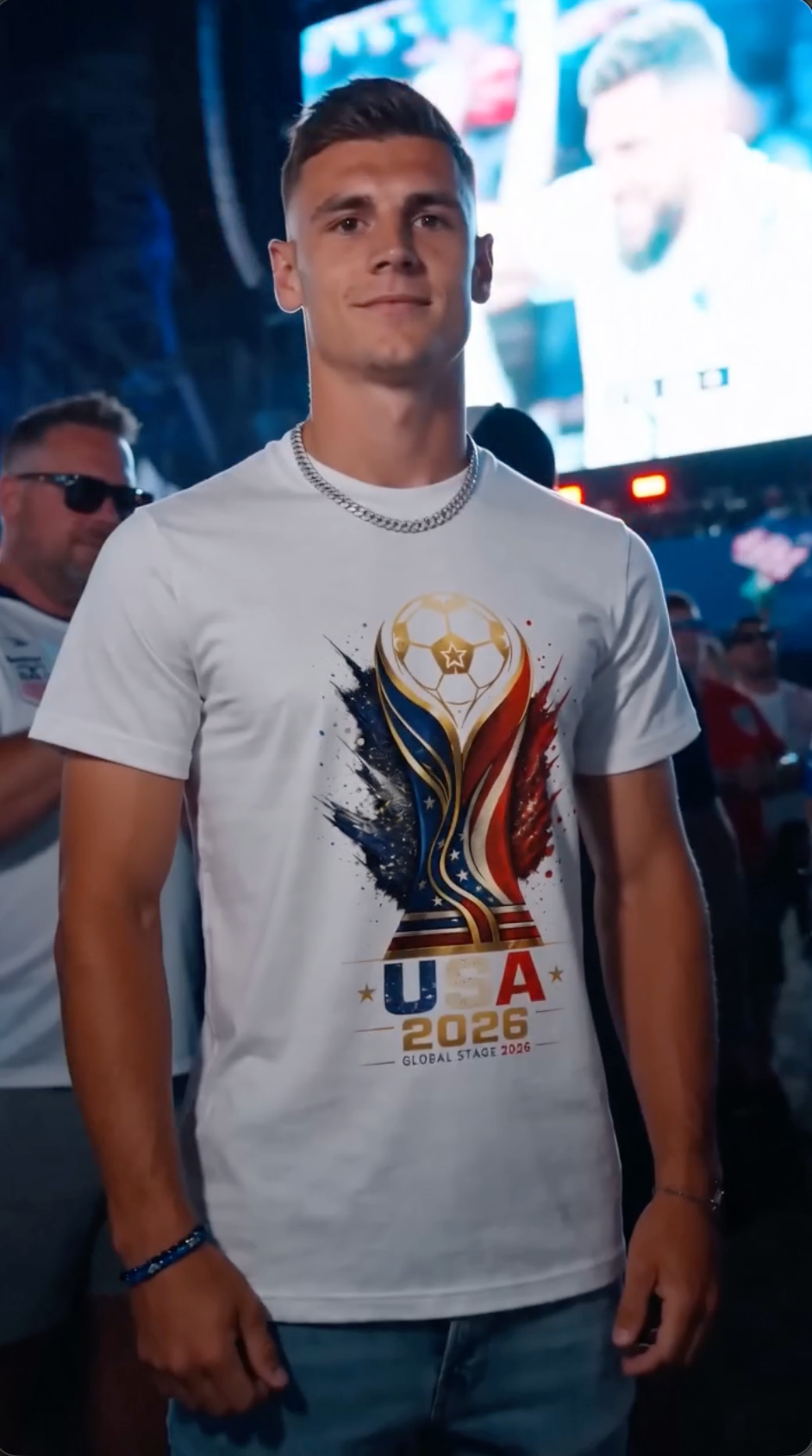 Male soccer fan wearing white USA 2026 Global Stage graphic t-shirt with patriotic soccer trophy design at a nighttime stadium event and fan celebration