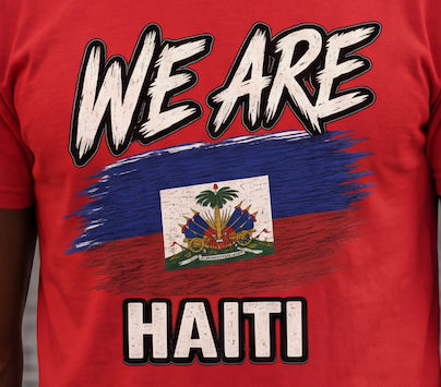 A close-up of a red t-shirt featuring a graphic print with the words 'WE ARE' and 'HAITI' in white distressed lettering, surrounding a Haitian flag design with the national coat of arms.