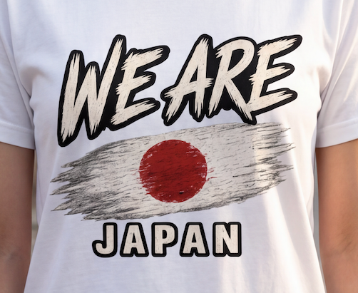 A close-up of a white short-sleeve t-shirt worn by a person, featuring a graphic print with the text 'WE ARE' and 'JAPAN' in a distressed brush font, surrounding a stylized Japanese flag design.