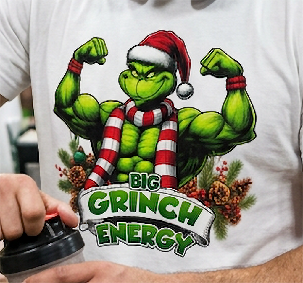 Close-up of muscular Grinch Christmas graphic on white t-shirt featuring Big Grinch Energy lettering and colorful holiday decorations.