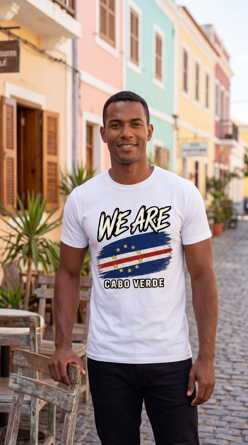 A man wearing a white graphic t-shirt with 'WE ARE CABO VERDE' and a flag design, standing on a cobblestone street in front of colorful pastel buildings.