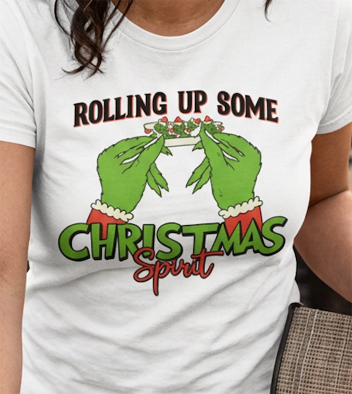 Close-up of white stoner Christmas t-shirt showing green hands rolling a holiday joint with bold Christmas lettering.