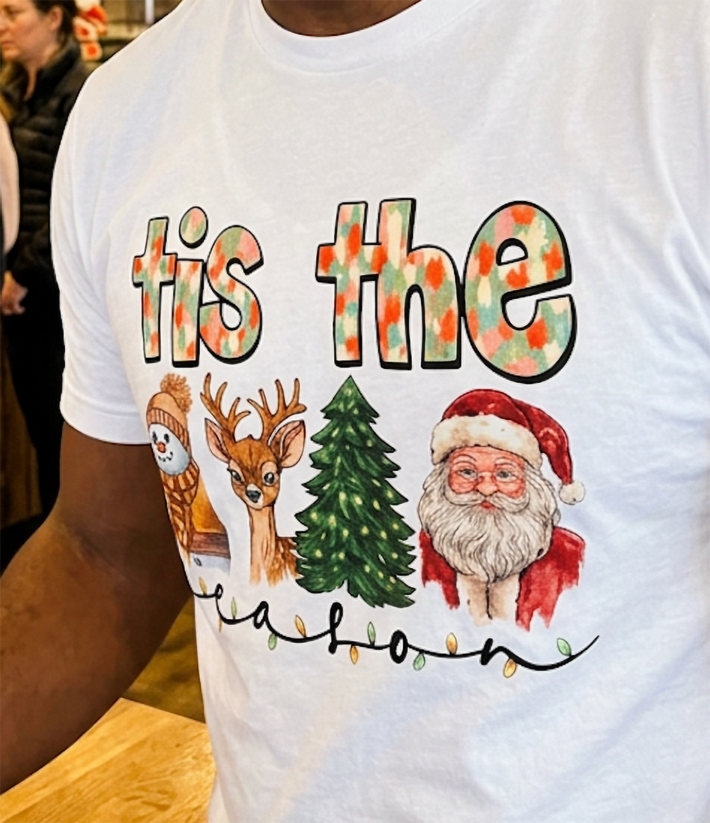 Close-up of white Tis the Season Christmas shirt showing watercolor holiday characters, colorful lettering, and decorative Christmas lights.