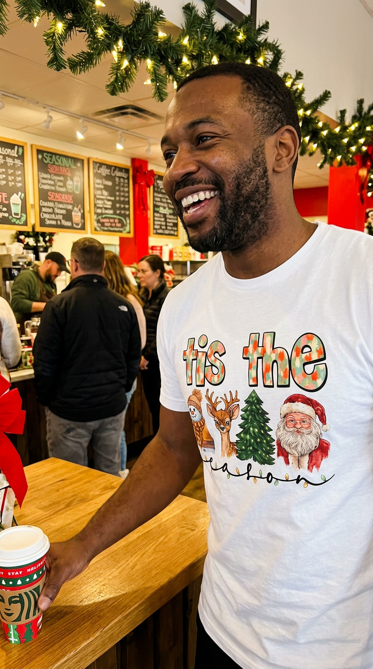 Man wearing white Tis the Season Christmas graphic tee while standing inside a festive store decorated for the holiday season.