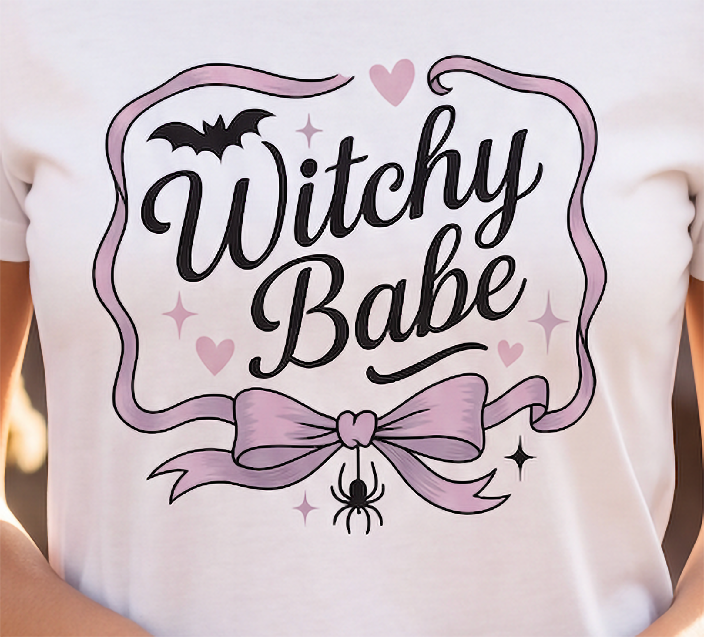 Close-up of pink and black Witchy Babe graphic on white Halloween T-shirt with bow, bat and spider details