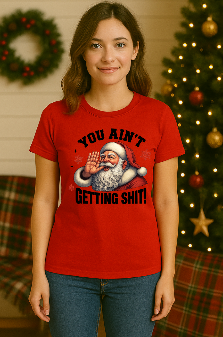 Woman wearing a red funny Christmas t-shirt featuring a Santa Claus graphic with the phrase “You Ain’t Getting Shit,” standing in front of a decorated Christmas tree.