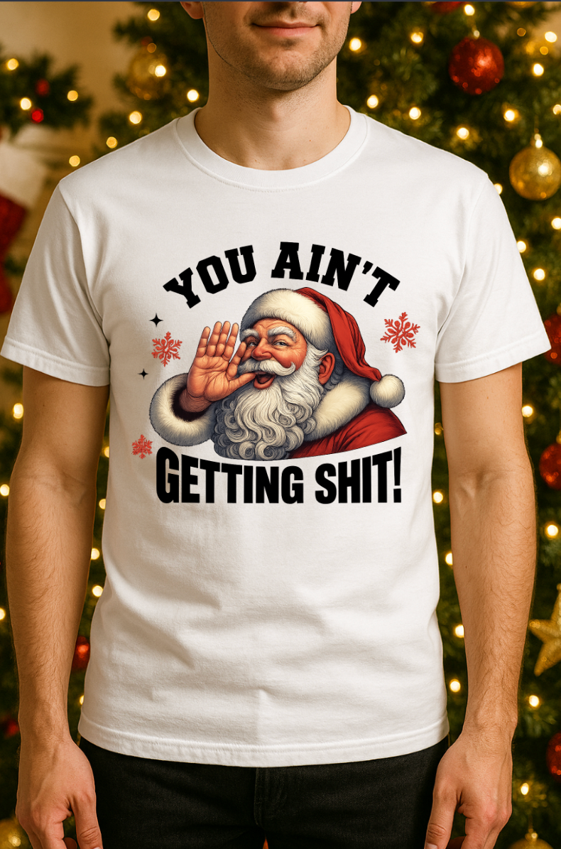 Man wearing a white funny Christmas t-shirt featuring a Santa Claus graphic with the phrase “You Ain’t Getting Shit,” standing in front of a decorated Christmas tree.