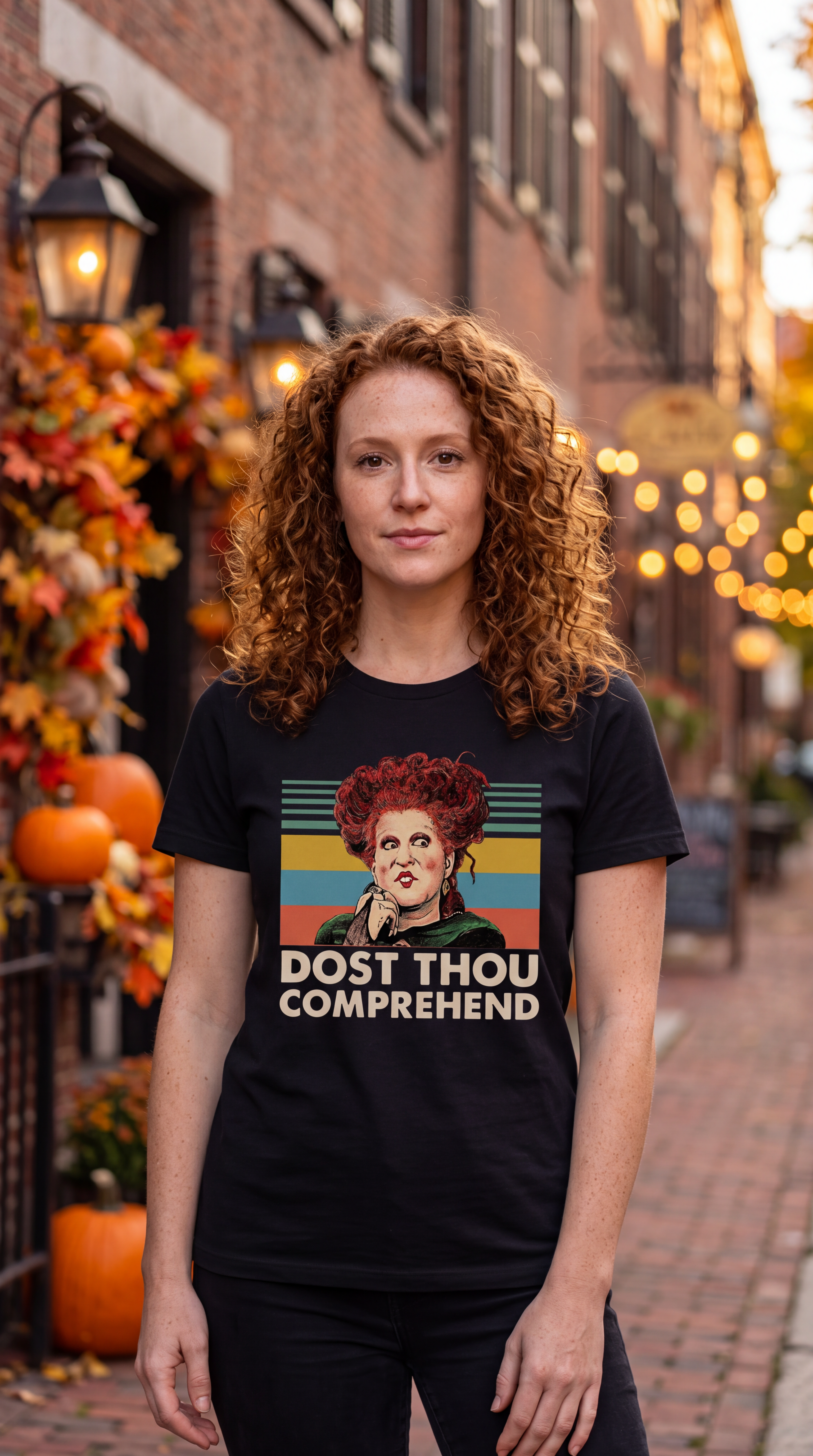 A woman with curly red hair stands on a brick street wearing a black graphic T-shirt featuring a retro-style illustration of a woman with red hair and the text 'DOST THOU COMPREHEND'. The background shows a cozy, autumnal street scene with pumpkins, fall leaves, and warm string lights.