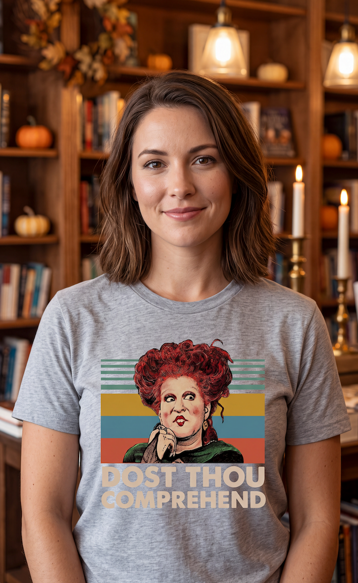 A woman with shoulder-length brown hair stands in front of a warm, book-filled background wearing a heather gray t-shirt with a colorful graphic of Red Riding Hood and the text 'DOST THOU COMPREHEND'.